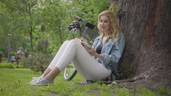 Portrait Cute Smiling Woman Sitting Under an Old Tree in the Park Reading a Journal Aloud. The