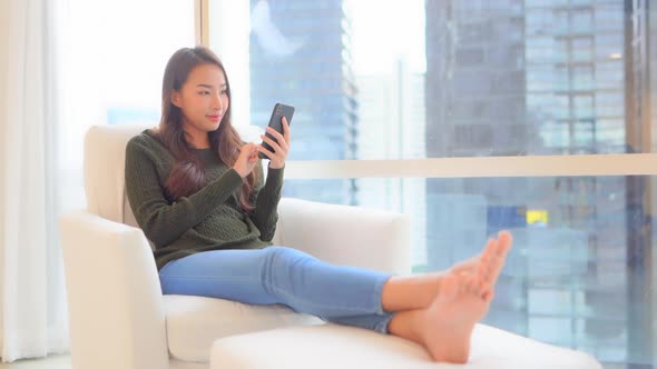 Asian woman sitting by skyscraper panoramic window with city view using smartphone
