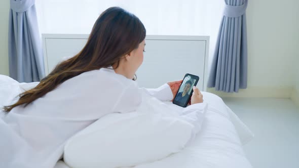 Asian beautiful woman in pajamas enjoy talk on video call on phone with boyfriend on bed at home.