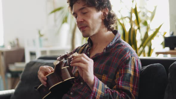 Concentrated adult man in a plaid shirt tunes the ukulele