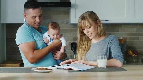 Young Family in the Kitchen. Mom Reading a Book, with the Baby Daddy Is Near