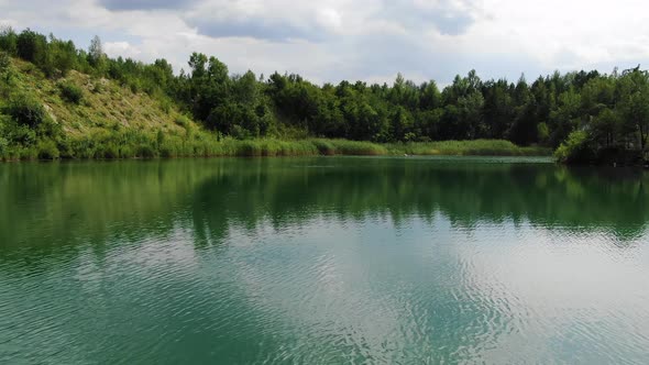 Low View of Calm Summer Lake With Grass in Foreground Surrounded by Forest Slowly Tracking Forward