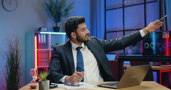 Businessman Sitting in front of Camera in Office and Explaining Records on Glass Wall, Gesturing