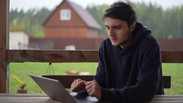 A Young Man is Typing on a Laptop While Sitting on a Terrace Against the Backdrop of a Picturesque