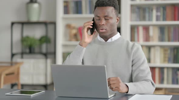 African Man Talking on Smartphone While Using Laptop in Library