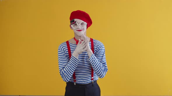 A greedy mime man wearing white makeup, striped shirt and red hat and suspenders rubbing his hands t