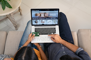  website with laptop at home. High angle view of man and woman using laptop on legs and paying with credit card. Indian casual couple selecting product on ecommerce portal on computer.