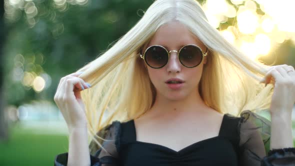 Cute Young Caucasian Blonde Girl in Round Stylish Sunglasses and a Black Dress Effectively Fluffs