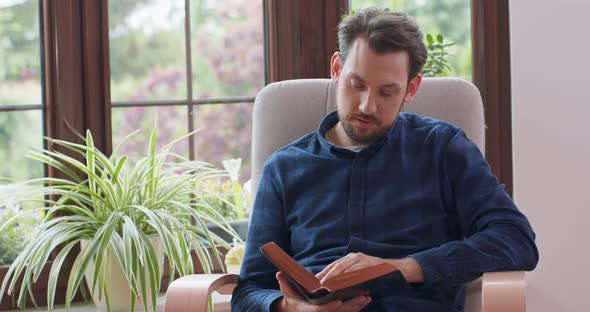 Young Bearded Man Reading Book While Sitting in White Chair Young Man Relaxing at Home Window and