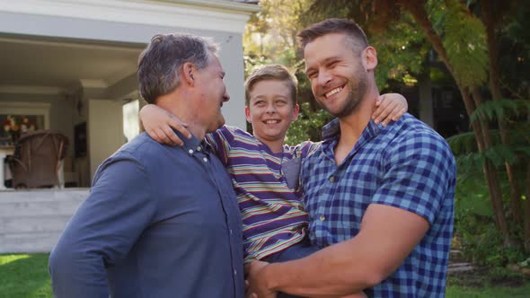 Portrait of happy caucasian grandfather, grandson and father embracing in garden and smiling