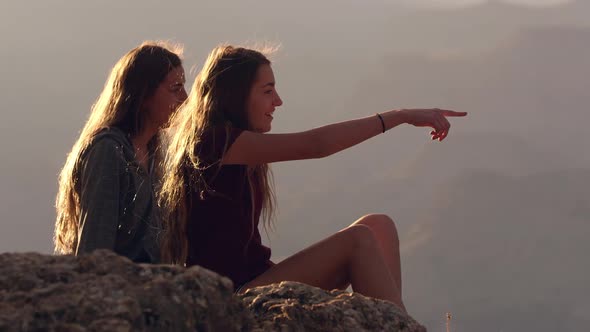 Twin teenage girls talking and pointing sitting on rocky edge