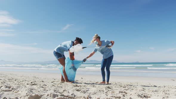 Two diverse women wearing volunteer t shirts and face masks picking up rubbish from beach