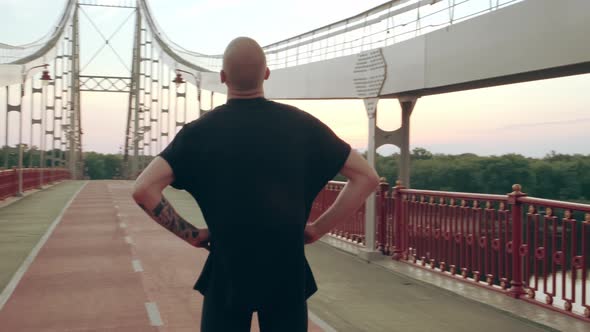 Young White Man in Black Sport Uniform Doing Warmup Neck Before Run Pedestrian Bridge at Dawn