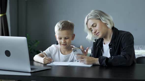 Mother Helping Son With Homework Sitting At Desk In Bedroom. The Child Does Homework