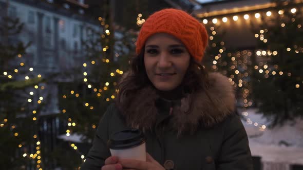 Portrait of an Attractive Girl in a Bright Hat with Coffee in Hands in the Winter on a Background of