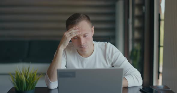 Concentrated Young Man Sits at Desk in Room with Laptop