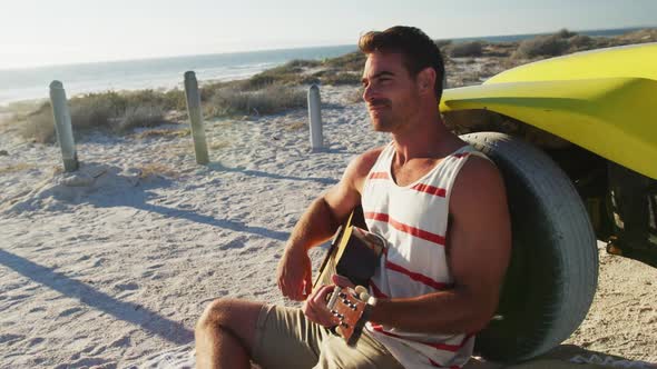 Happy caucasian man sitting beside beach buggy by the sea playing guitar