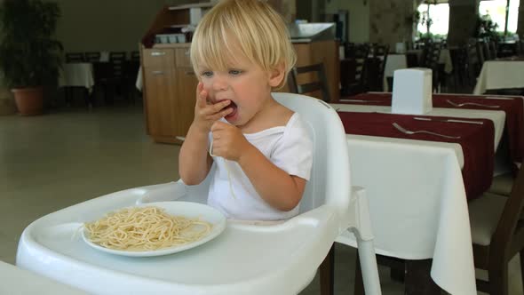 Baby Boy Eats Spaghetti Sitting in a Baby Chair in Restaurant