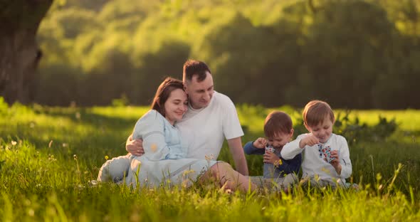 Loving Parents Hugging Sitting in the Field at Sunset and Looking Smiling at Two Sons Eating Ice