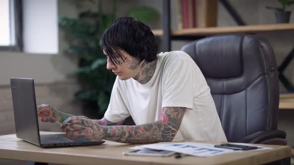 Confident Concentrated Young Man with Tattooed Hands and Face Messaging Online Typing on Laptop