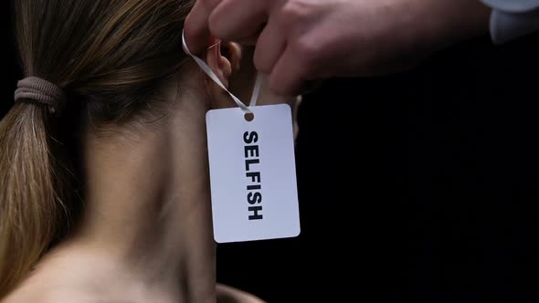 Man Hand Hanging Selfish Label on Female Ear, Showing Disrespect and Shaming