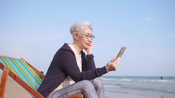 Asian elderly woman chatting with friends on video call while relaxing at the beach.