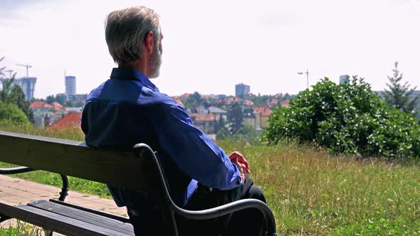 Old Senior Man Sits on the Bench in the Park and Looks at the City