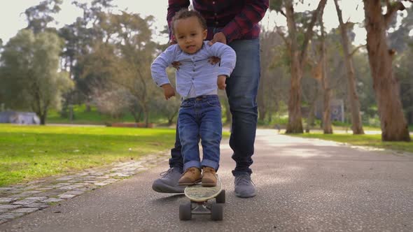 Little Boy Standing on Skateboard, Rolling with Help of Father 