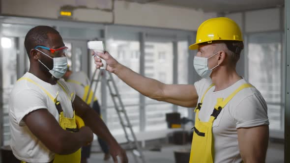 Foreman in Uniform and Safety Mask Measuring Temperature of Builders with Contactless Thermometer