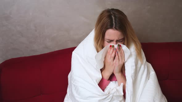 Young Caucasian Ill Girl with Covid or Flu Sitting Under Blanket on Couch in Living Room and Blowing