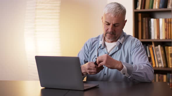 Mature Man Wiping and Putting on Glasses and Working with Laptop Computer at Home Office