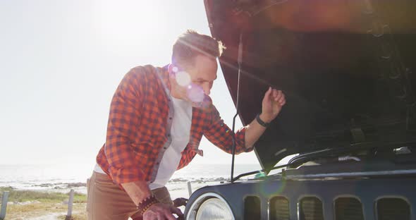 Stressed caucasian man looking at broken down car with open bonnet on sunny day at the beach