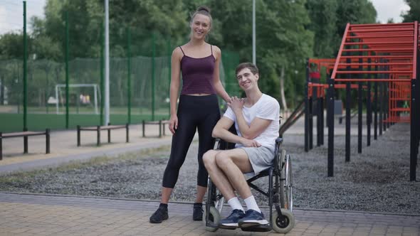 Wide Shot of Smiling Young Woman Posing with Man in Wheelchair on Sports Ground