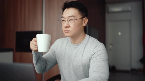 Asian serious man wearing eyeglasses working on laptop and drinking coffee