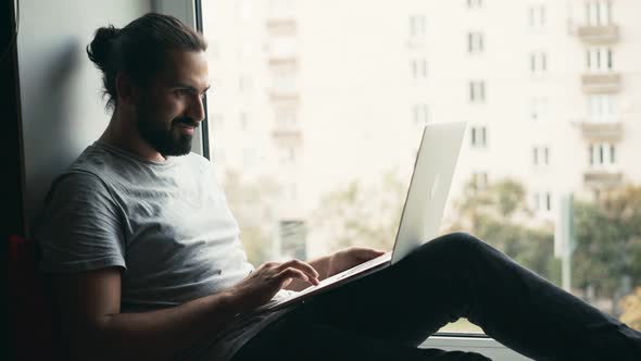 Young Handsome Man Working on His Laptop While Sitting on a Windowsill