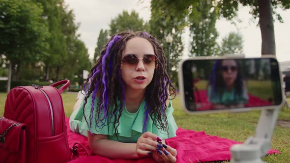 A Woman with a Stylish Hairstyle Lies on a Blanket in the Park and Records a Video Blog Using Her