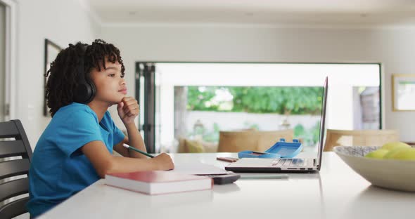 Happy biracial boy sitting at table using laptop and having online classes