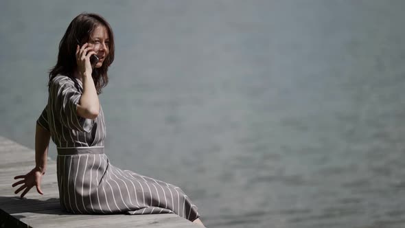 a Brunette in a Striped Dress is Sitting on a Wooden Deck By the River on a Sunny Day