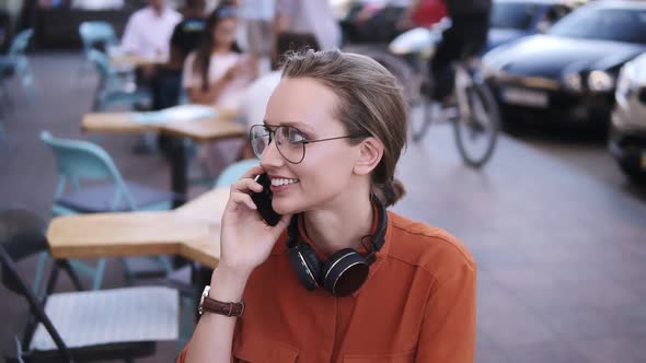 Close Up of Fair Hair Girl with Headphones on Her Neck Talking on Mobile Phone While Sitting in