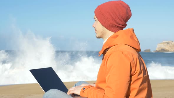 Man Sits on Ocean Beach with Laptop and Enjoys Distance Work