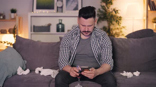 Sensitive Guy Crying Holding Glass of Wine Using Smartphone Sitting at Home