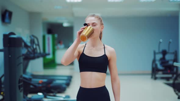 Young Woman in Black Sportswear Walks Through the Gym with a Shaker in Her Hands 