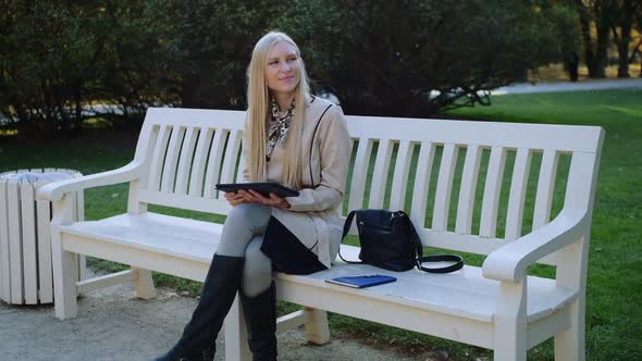 Young Beautiful Woman Sits On The Bench In Autumn Park And Works On The Tablet