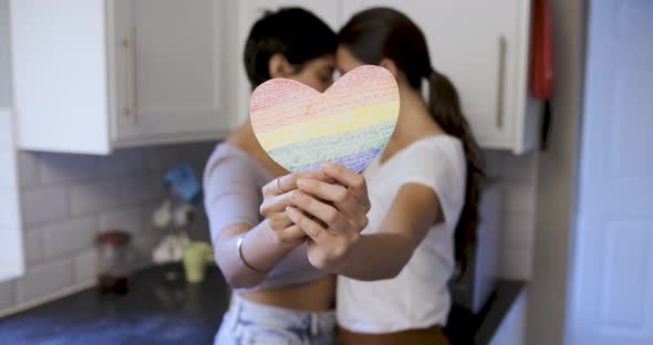 Happy girl friends lesbian couple embracing and holding a heart shaped rainbow flag