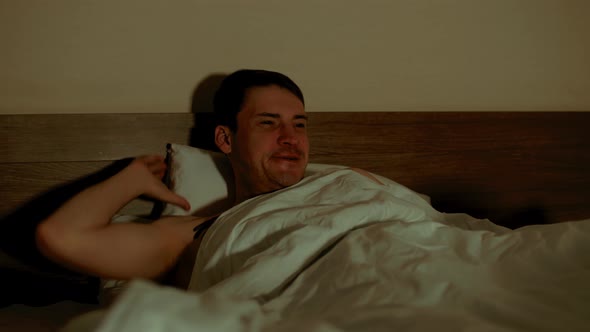 Young Man Lying on Pillow Under Blanket in Bed and Watching Tv in Late Evening