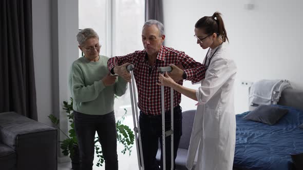 Old Man with Crutches Making First Steps with Support of Wife and Doctor