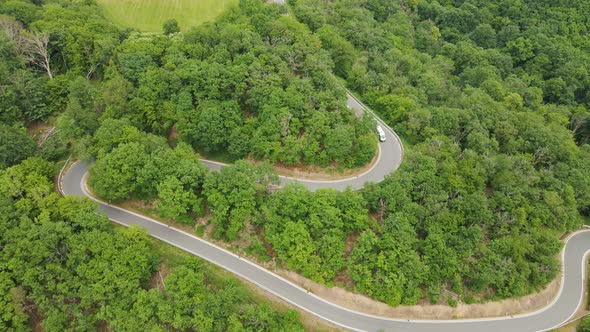 A silver recreational vehicle driving down an empty hillside road with several hairpin bends. Aerial