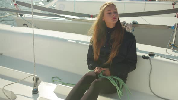 A Girl with Long Hair Sitting on the Deck of a White Sports Yacht Unwinds a Rope To Prepare the Boat