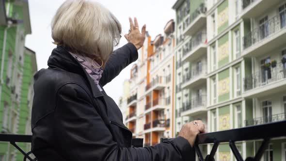 Side View Senior Woman Waving to Neighbors Standing on Urban City Balcony Outdoors