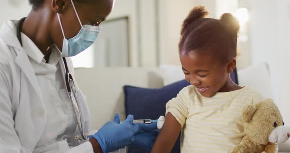 African american female doctor wearing face mask giving covid 19 vaccination to smiling girl at home